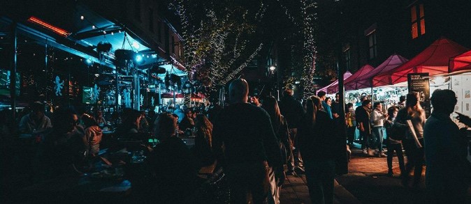 Crowds walking through a festive Christmas in July night market in Sydney, with illuminated stalls, street lights and outdoor dining areas.