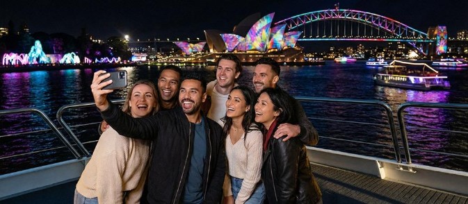 Group of friends taking a selfie on a Vivid Sydney cruise, with the illuminated Sydney Opera House and Harbour Bridge in the background at night.